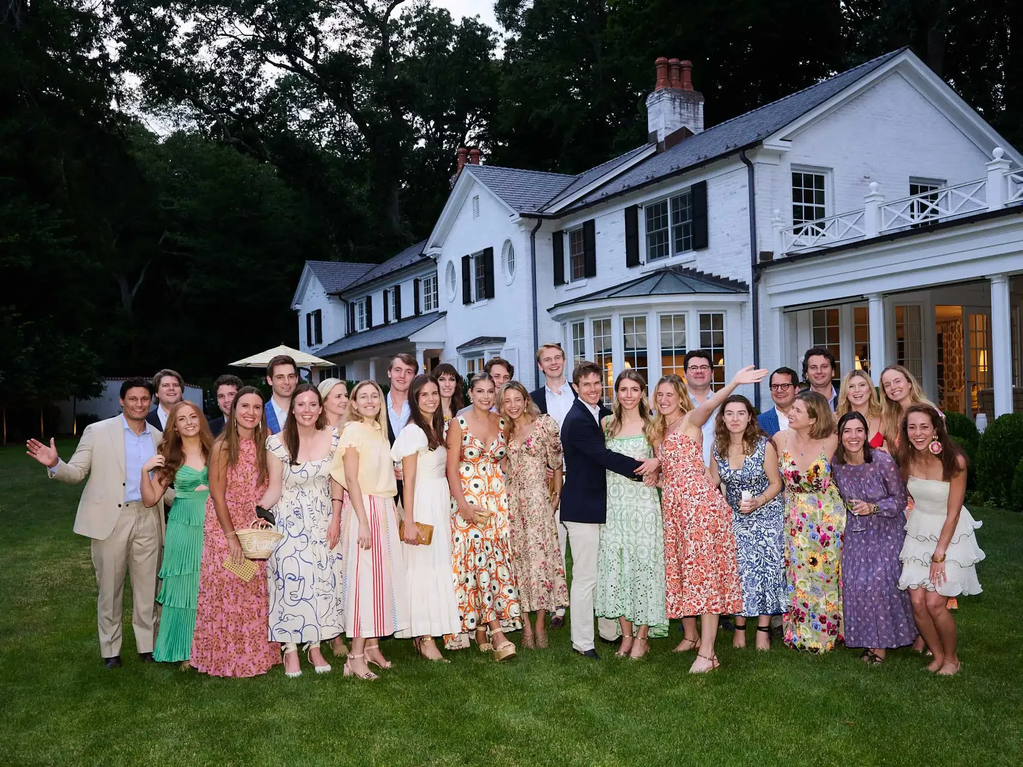Photo of a group of people in formal and summer dresses posing for a photo in front of a large white house with black shutters during evening.
