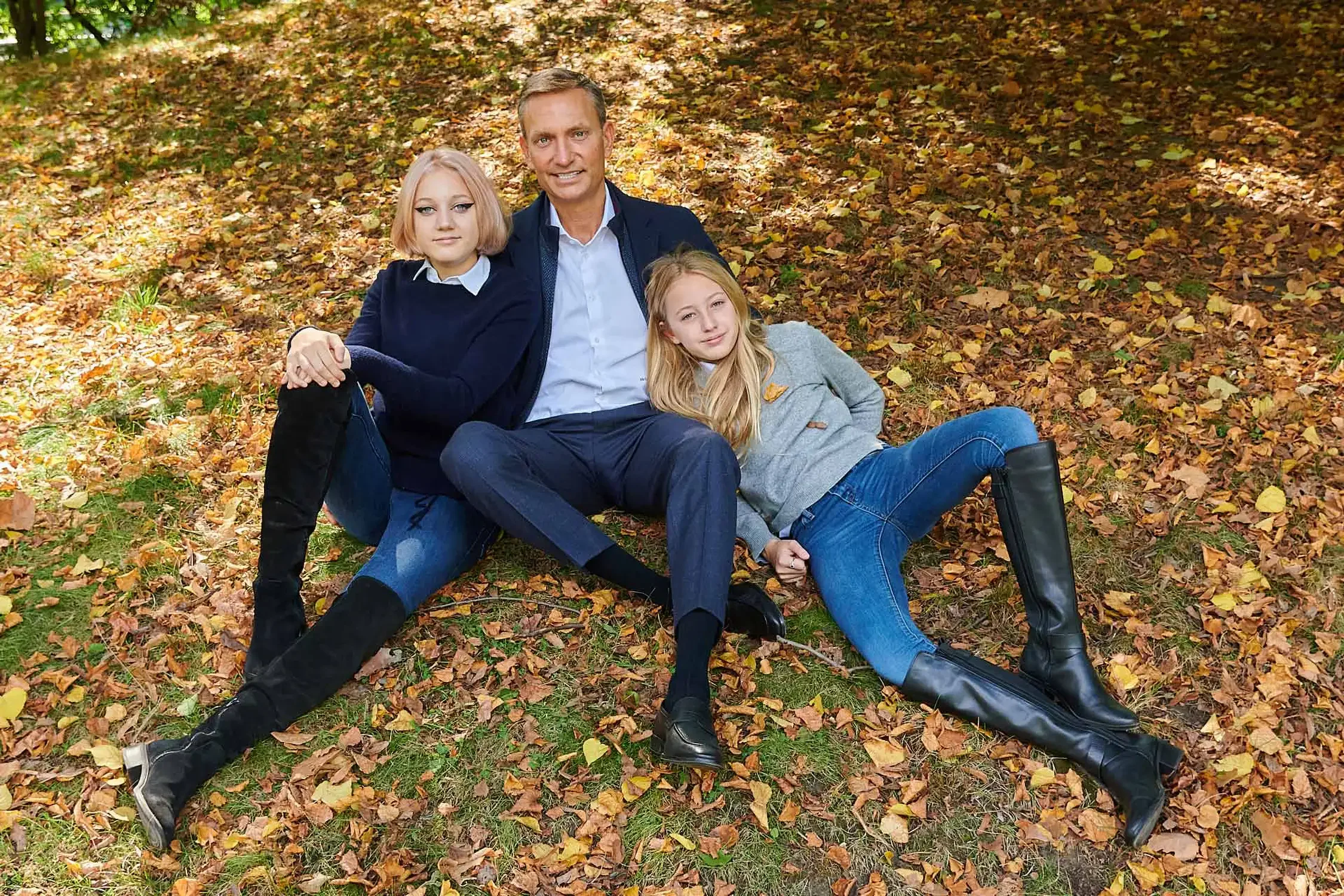 A man and his two young girls photographed  sitting on autumn leaves in a park. The father is in the middle, with one girl sitting to his left and the other girl lying on her side in front of him. All three are smiling and relaxed.