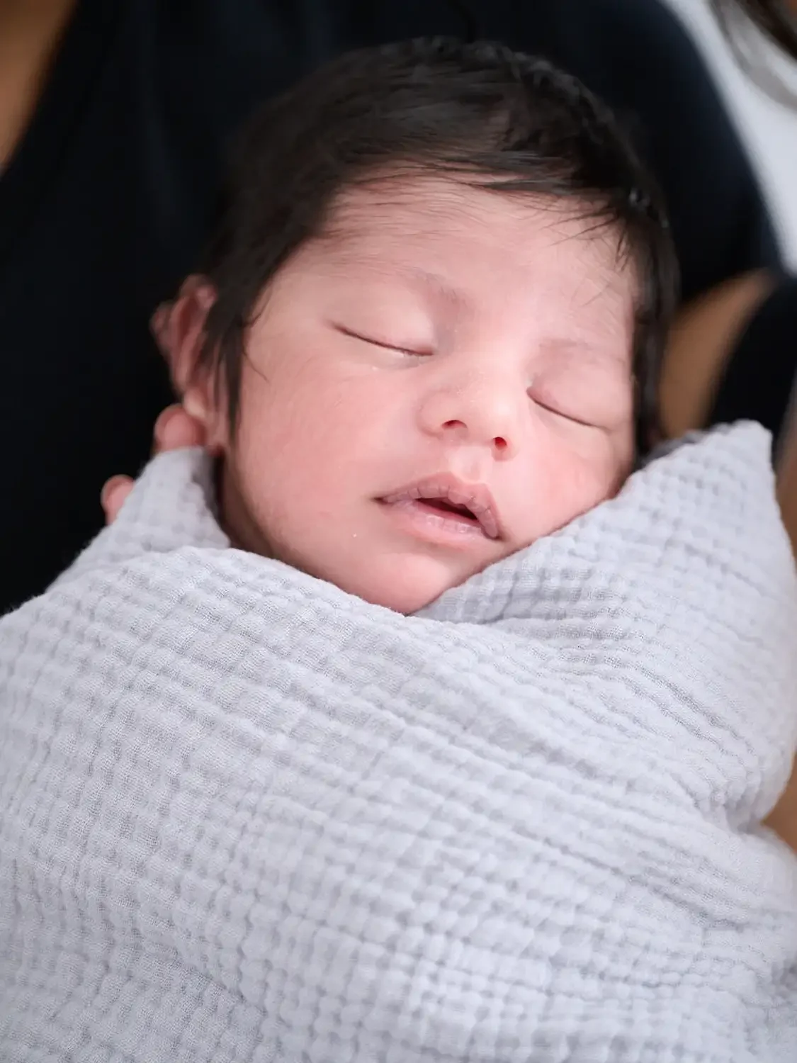 Family photo of a  sleeping newborn baby wrapped in a white textured blanket.