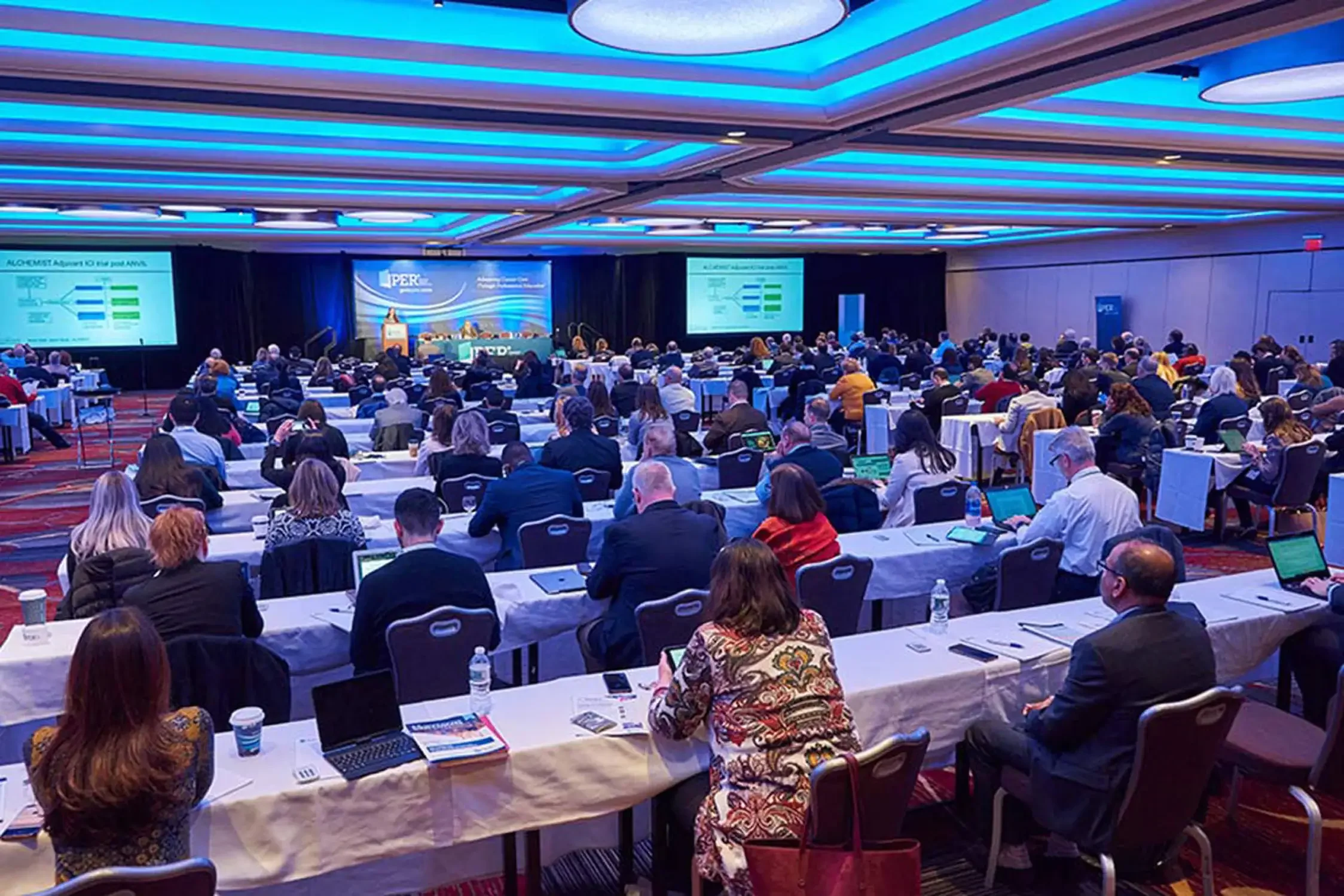 Corporate event photo of a conference room filled with attendees seated at long tables, facing a stage with large screens displaying presentation slides.
