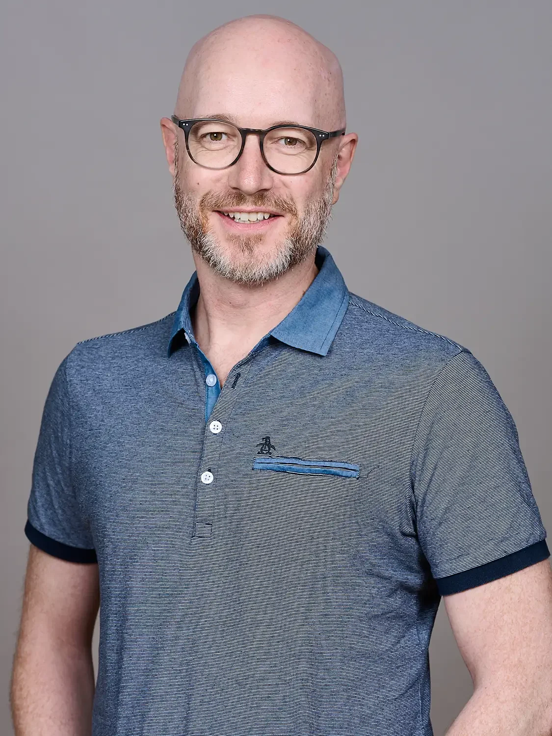 Headshot of a bald man with glasses and a beard, wearing a blue collared shirt, smiling against a neutral gray background photo studio setup.
