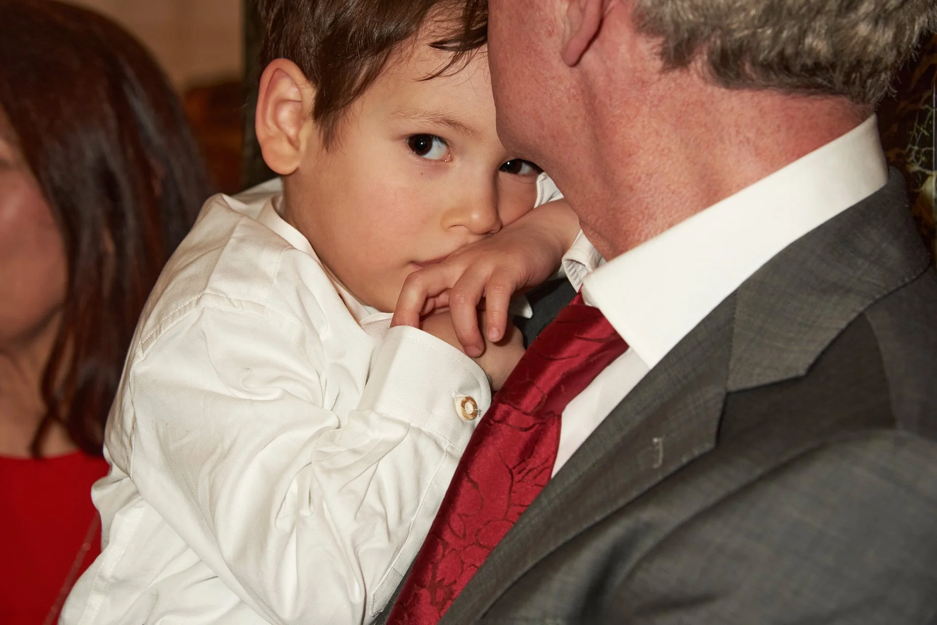 A young boy leaning on an older man's shoulder, resting his head against the man's face during a close moment. The boy has brown hair and is wearing a white shirt. The man is dressed in a suit and tie, with part of his face and white shirt collar vis