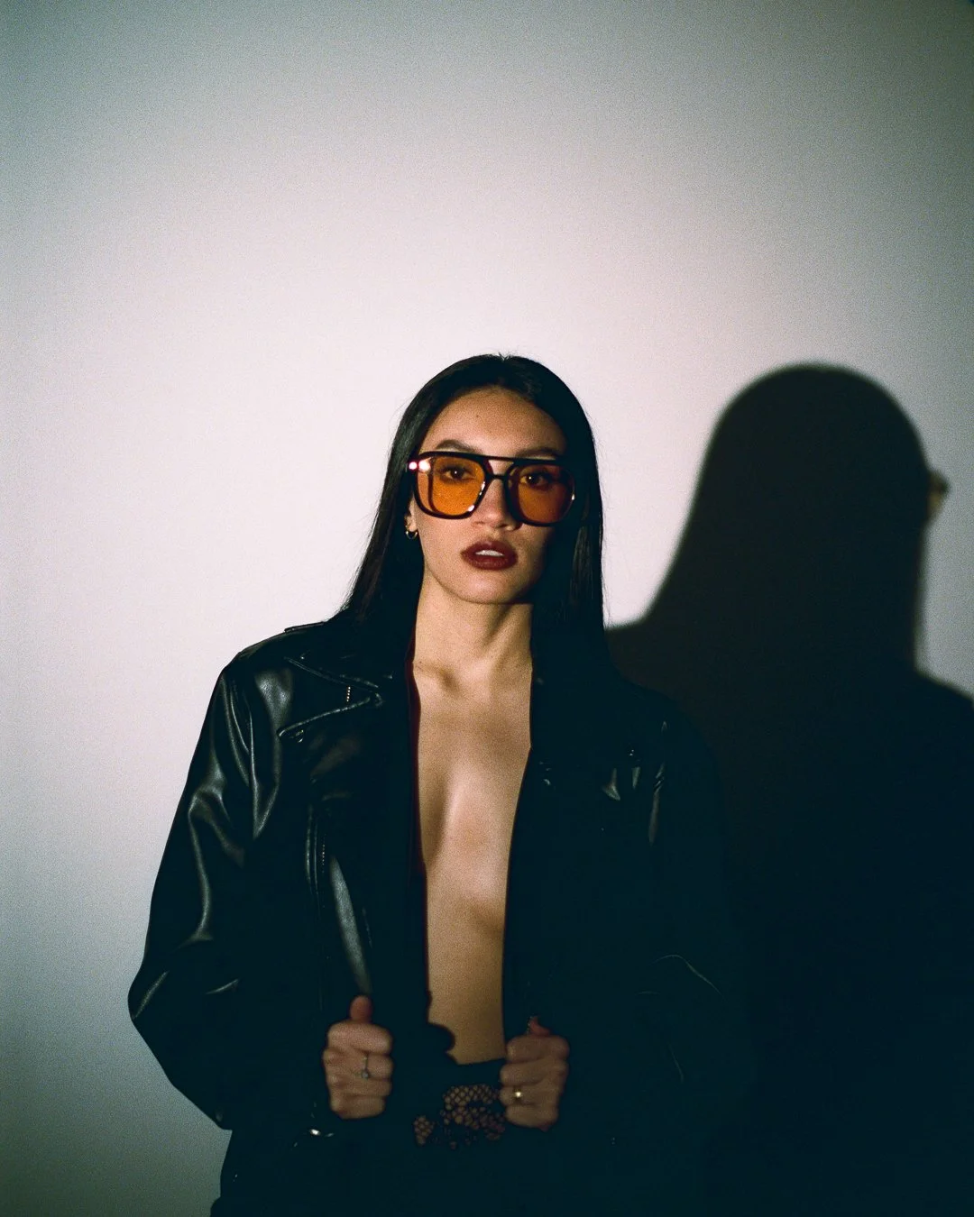 A woman with straight dark hair wearing oversized orange sunglasses, dark lipstick, a black leather jacket, and fishnet top, standing against a plain gray background with strong shadows.