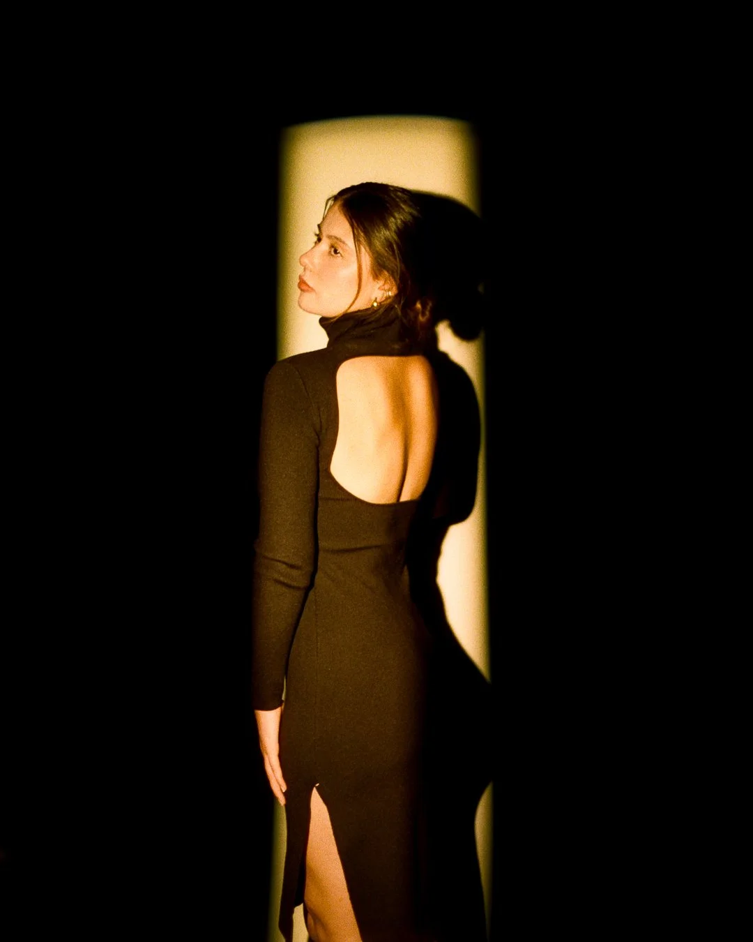 A woman in a black dress with an open back and slit standing in a spotlight, casting a shadow on the dark background.