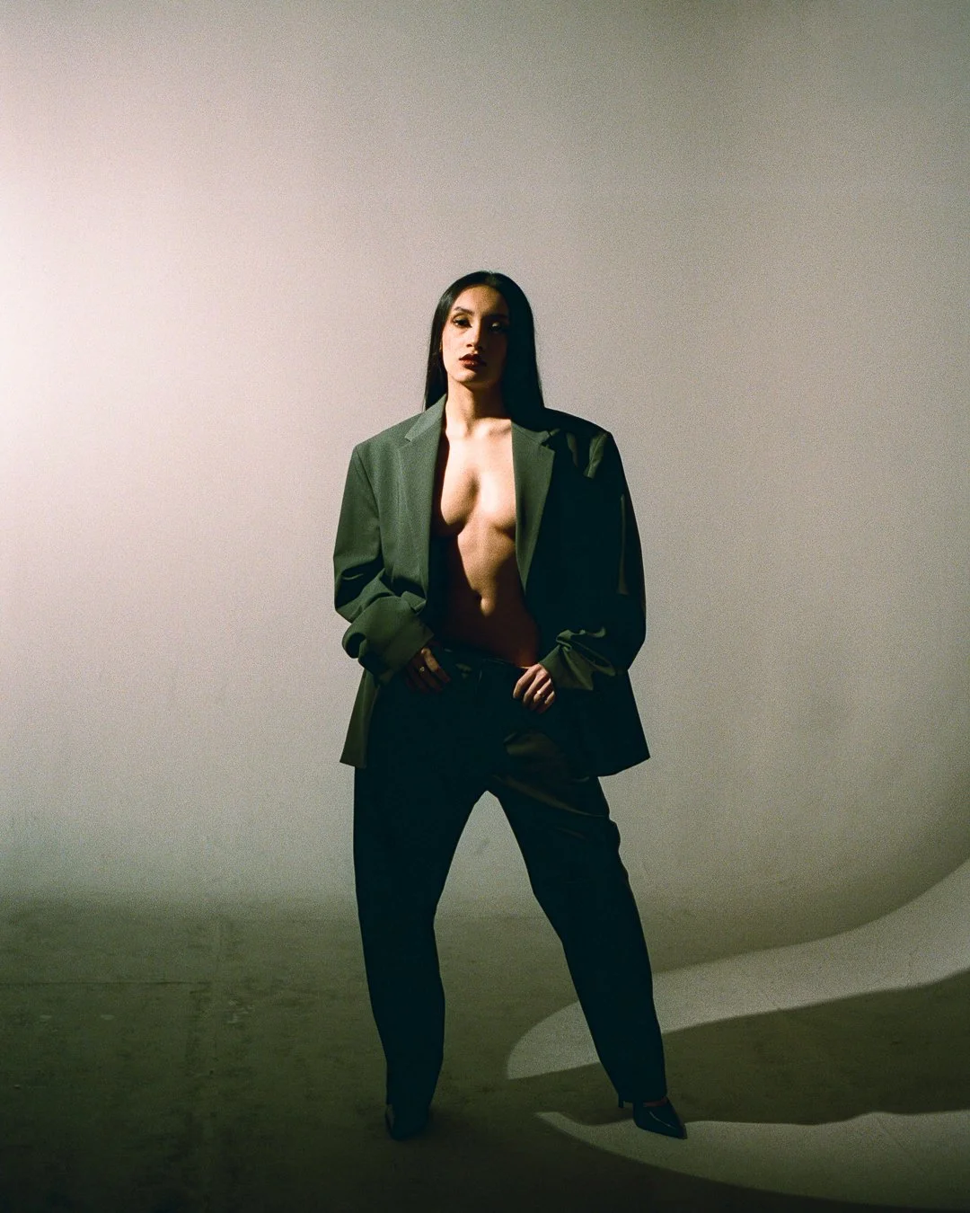 A woman with long dark hair stands confidently against a plain wall, wearing an oversized green blazer and black pants, with a serious expression.