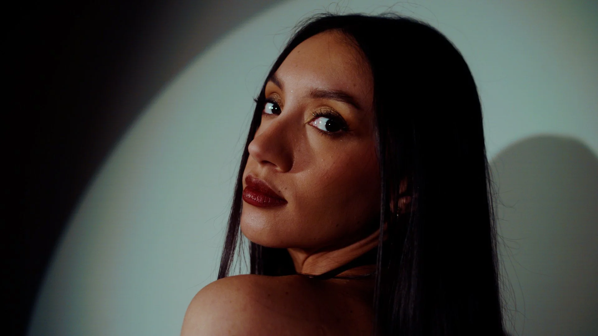 Close-up of a woman with long dark hair, makeup with gold eyeshadow, dark lipstick, looking over her shoulder at the camera, against a neutral background.