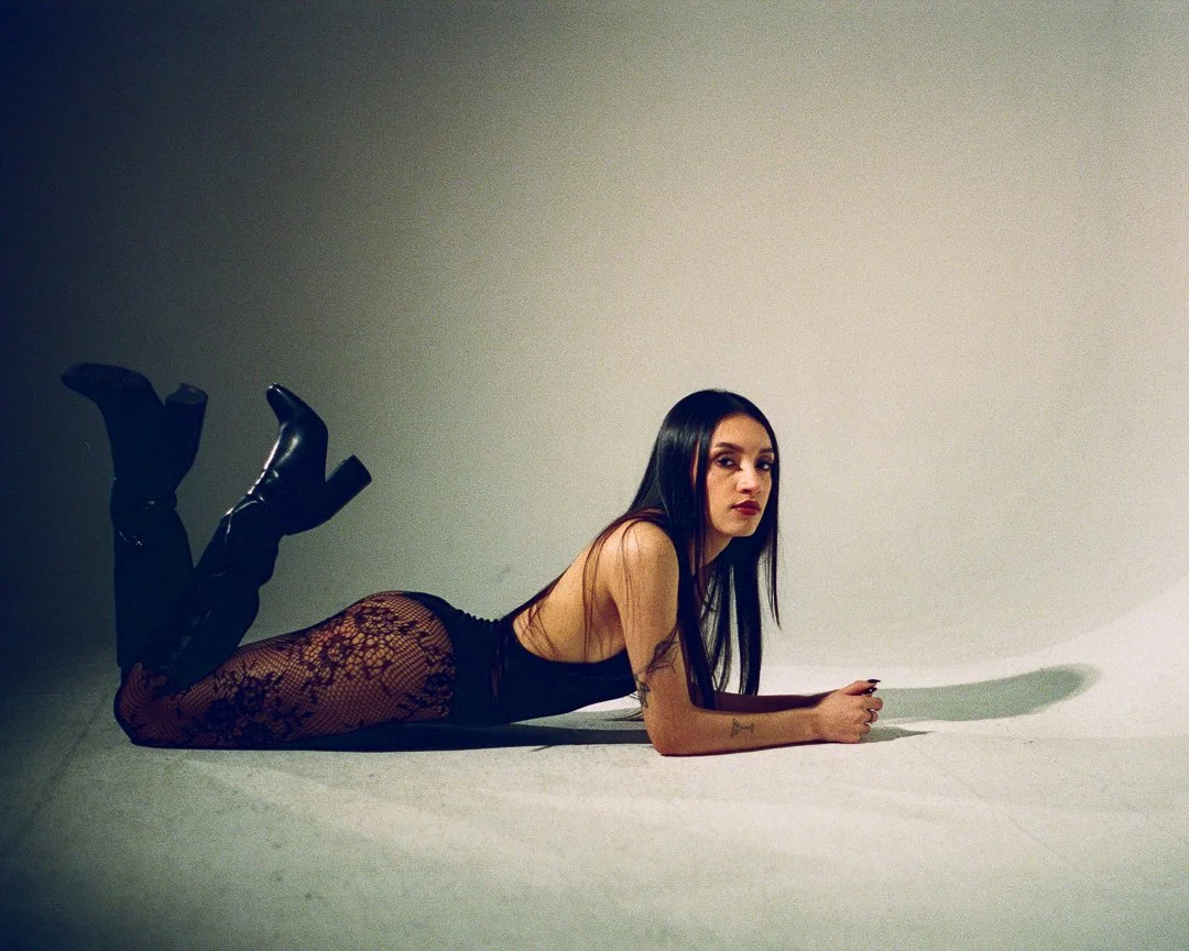 A woman lies on the floor facing the camera, wearing black lace tights, a black top, and black high-heeled boots, with long dark hair in a minimalistic setting.