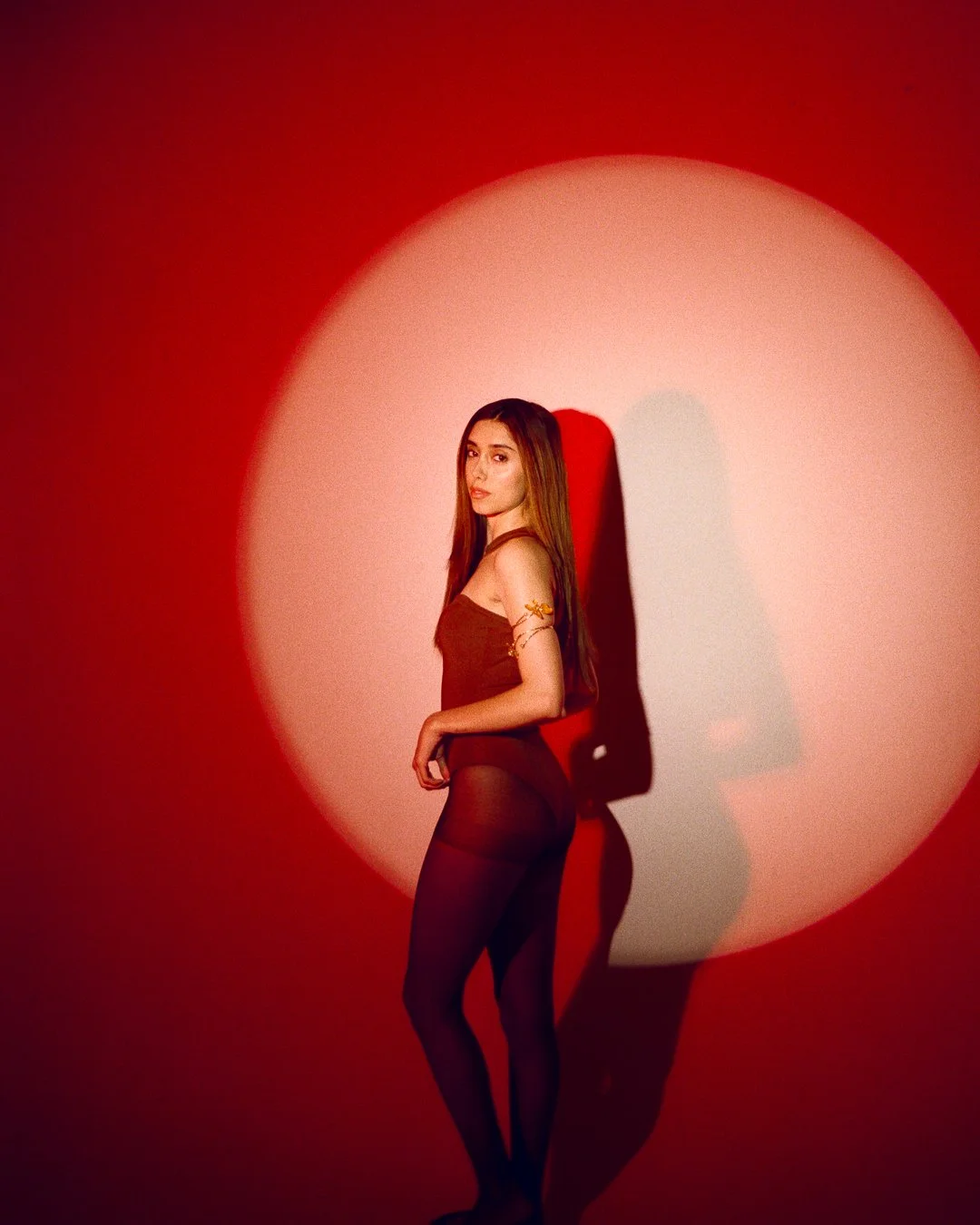 A young woman standing against a red wall with a large white circle spotlight. She is wearing a dark sleeveless top, sheer tights, and has a yellow floral tattoo on her arm. She is looking at the camera with a serious expression.