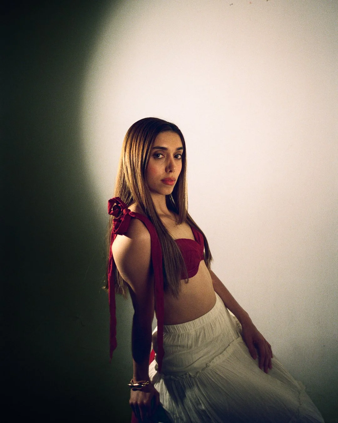A woman with long straight brown hair, wearing a red top with shoulder bows and white pants, sitting against a plain wall with a shadow cast on the wall.