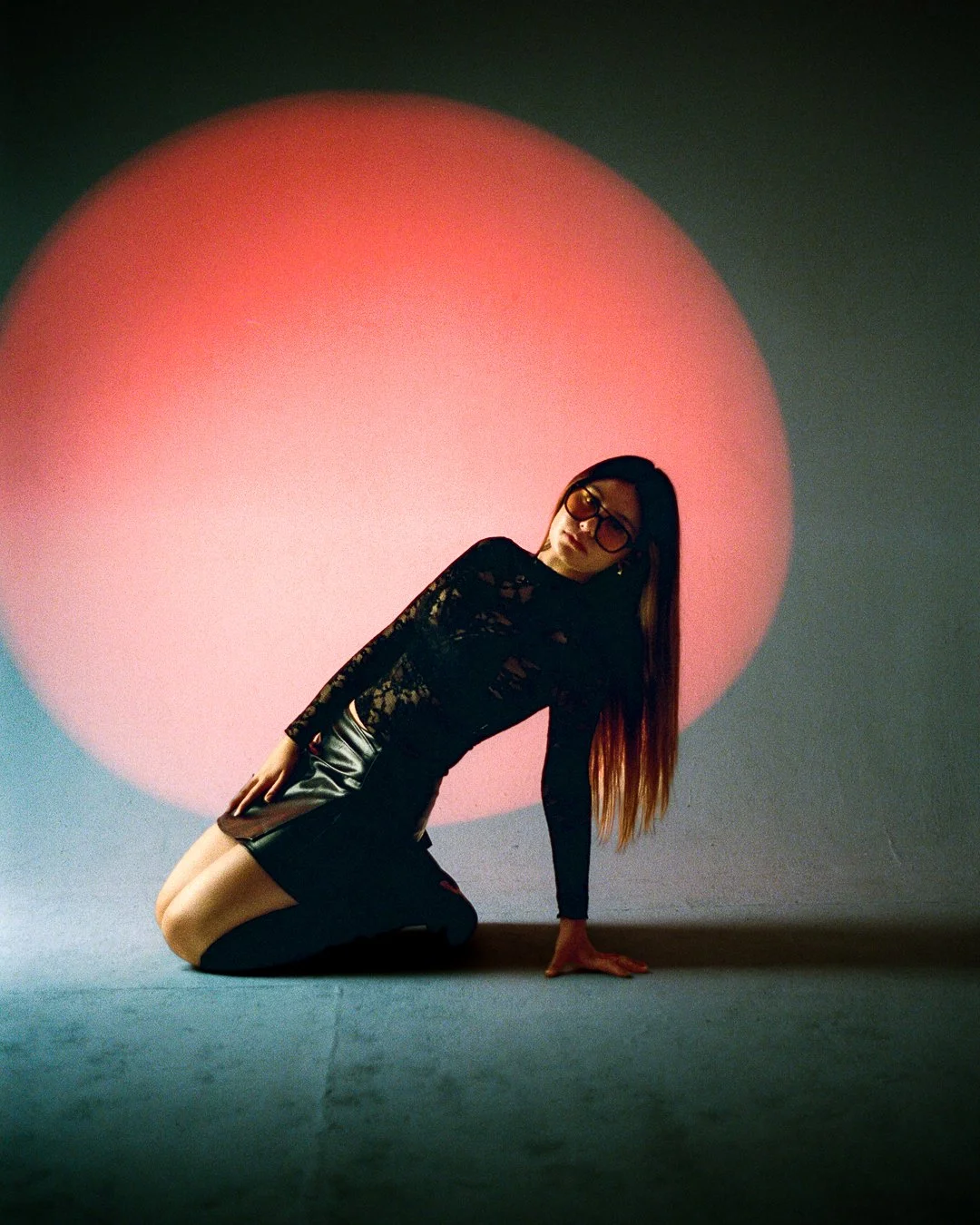 A woman with long hair, wearing glasses, a black lace top, and leather skirt, kneeling on the floor in front of a large red circular light projection.
