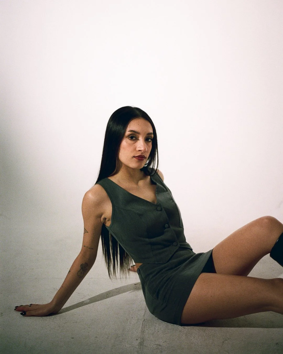 A woman with long dark hair sitting on the floor against a plain white wall, wearing a sleeveless dark green dress and black knee-high boots, with tattoos on her left arm.