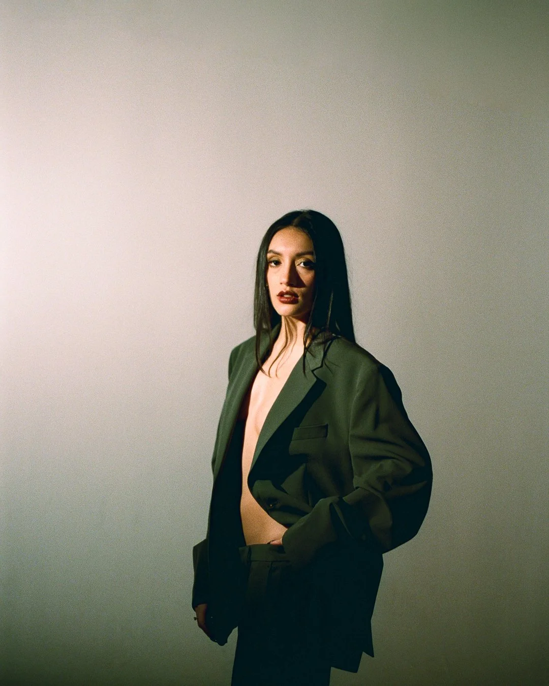 A woman with long dark hair wearing a dark green blazer and black pants, standing against a gradient grey background.