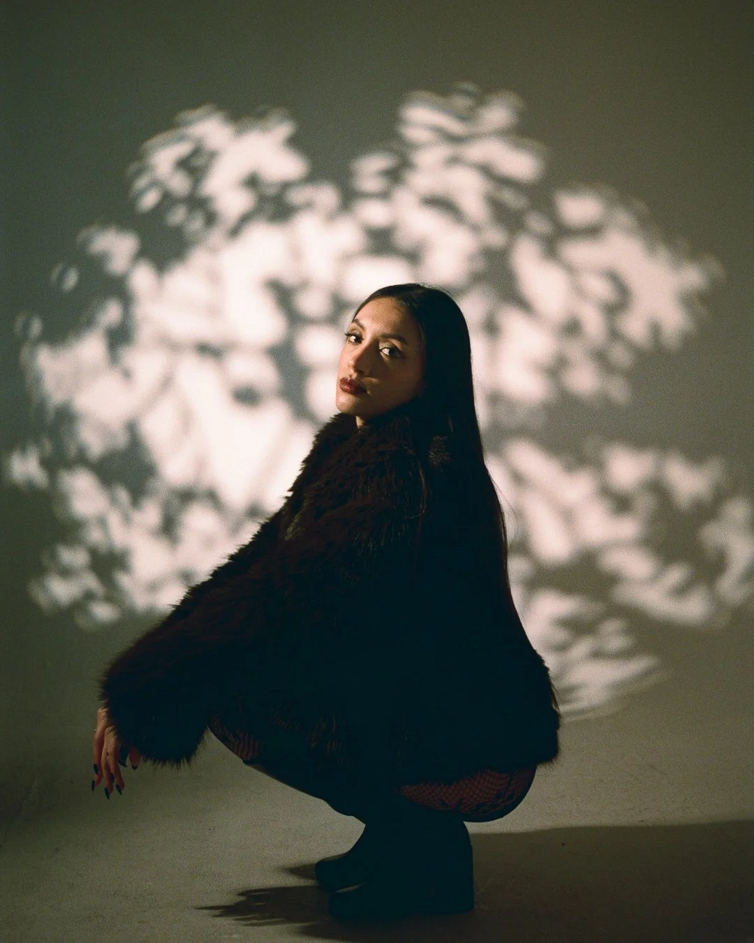A woman with long dark hair squats in a dark outfit, looking over her shoulder with a shadow of patterned light behind her on a neutral background.