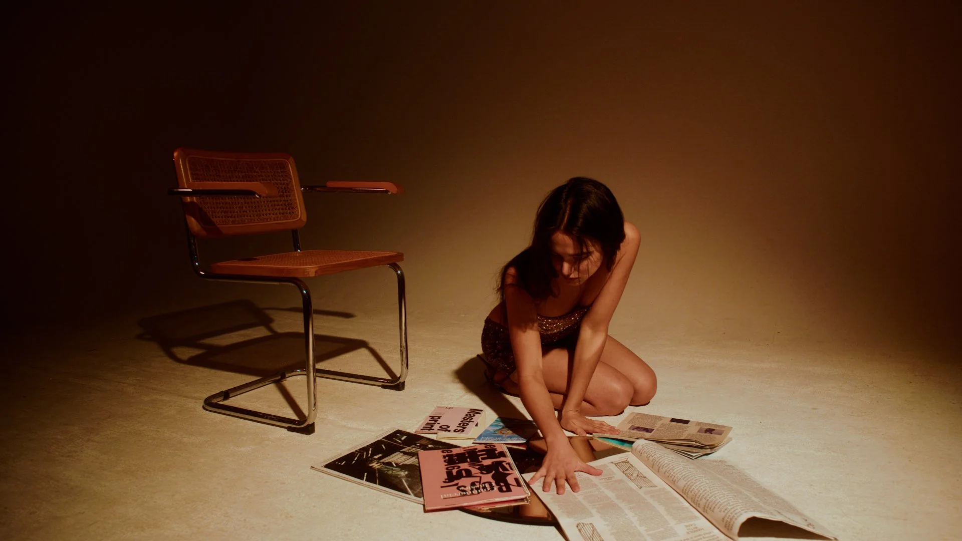 A woman with dark hair kneeling on the floor, reaching for open magazines and newspapers scattered around her, with an empty orange chair in the background under warm lighting.