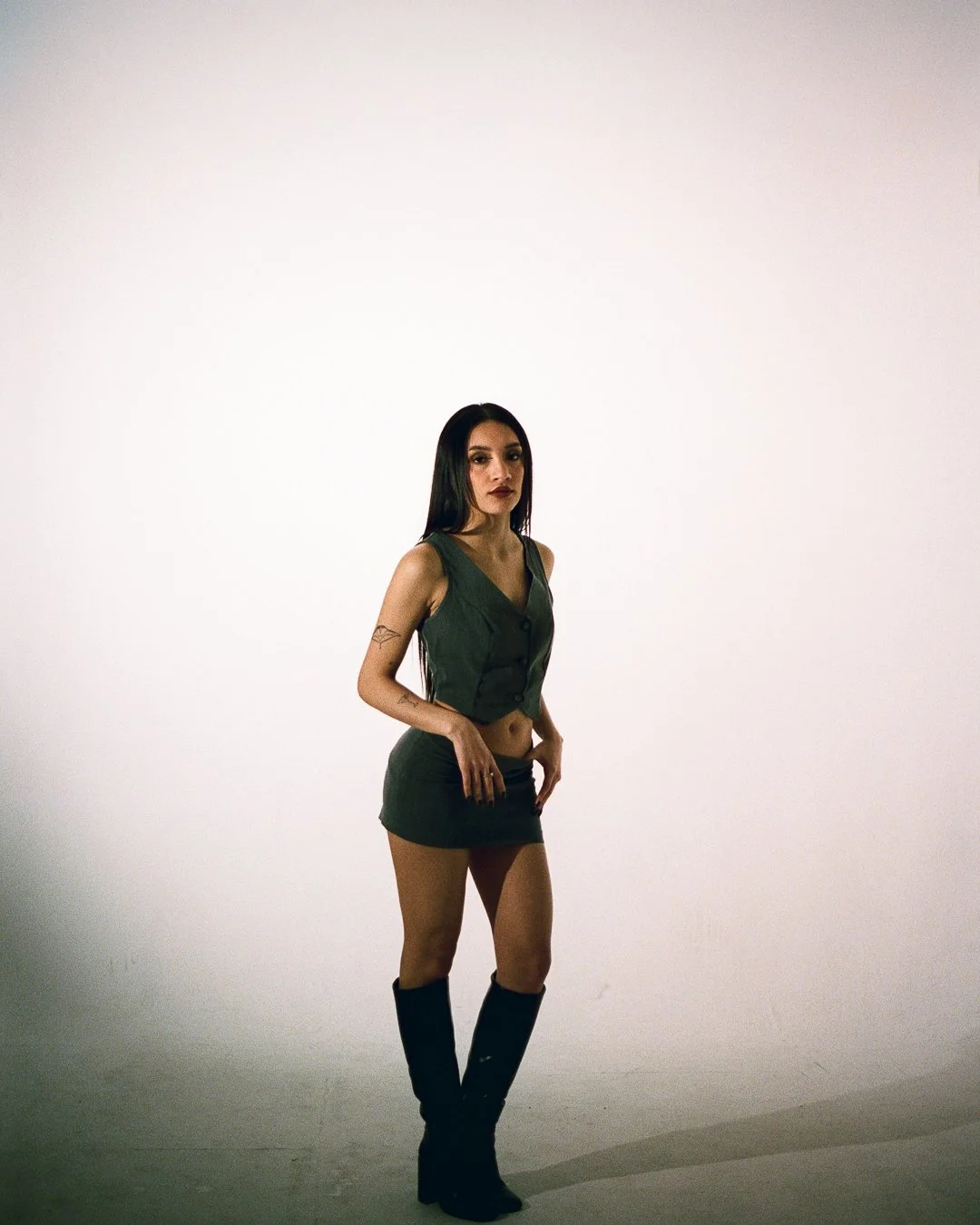 A woman with long dark hair and tattoos on her arm, wearing a sleeveless green vest, short skirt, and tall black boots, standing against a plain white background.