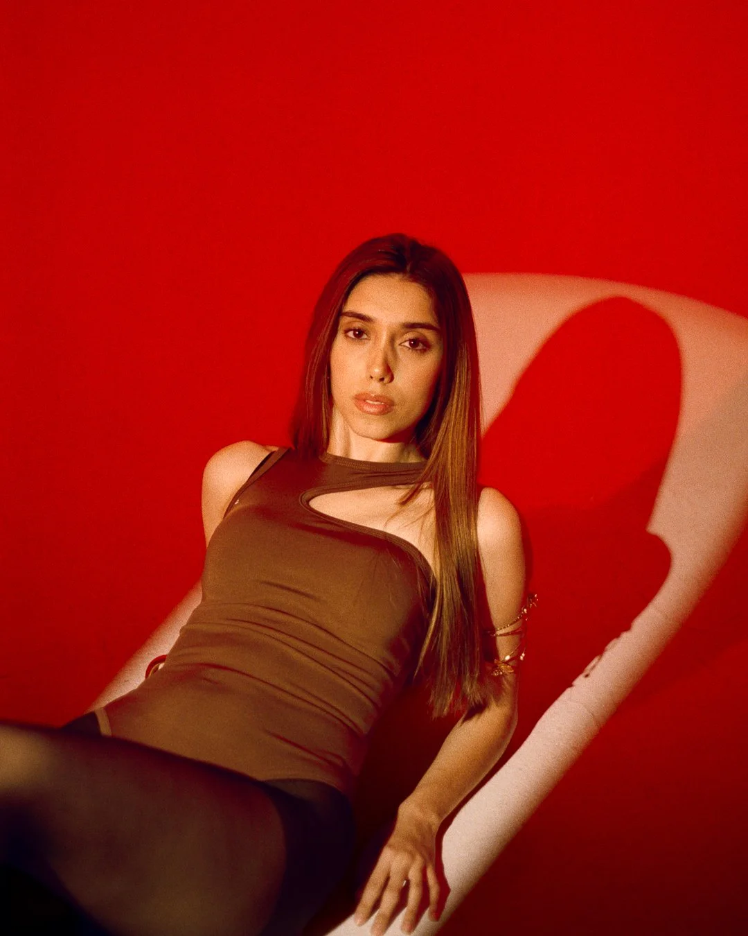 A woman with long brown hair, wearing a brown dress, sitting on a white lounge chair against a red background.