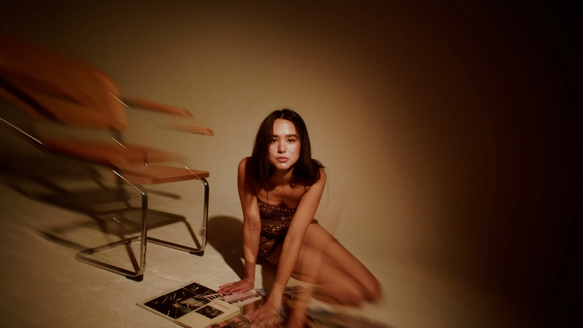 Young woman sitting on the floor in a dimly lit room with a beige wall, next to a stack of chairs and a magazine.