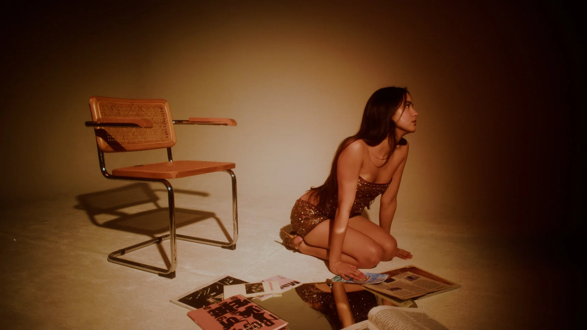 A woman in a sparkly dress kneeling on the floor with newspapers and magazines scattered in front of her, next to an empty wooden and metal chair, in a dimly lit room.