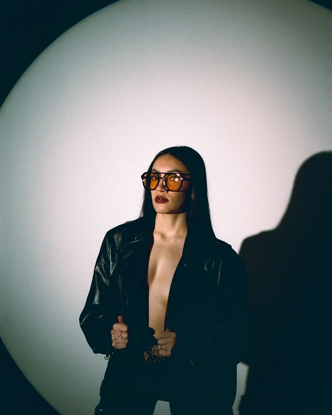A woman with long dark hair wearing large orange-tinted sunglasses and a black leather jacket standing against a plain white circular backdrop.