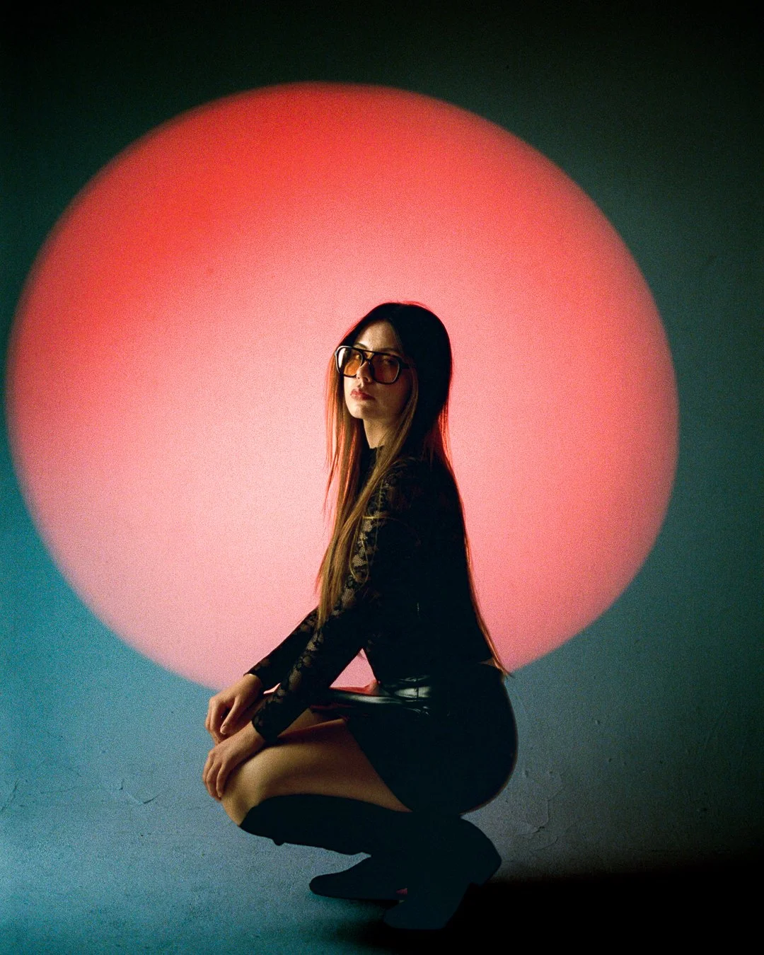 A woman with long hair, glasses, and a black outfit crouches in front of a large pink circle, against a dark background.