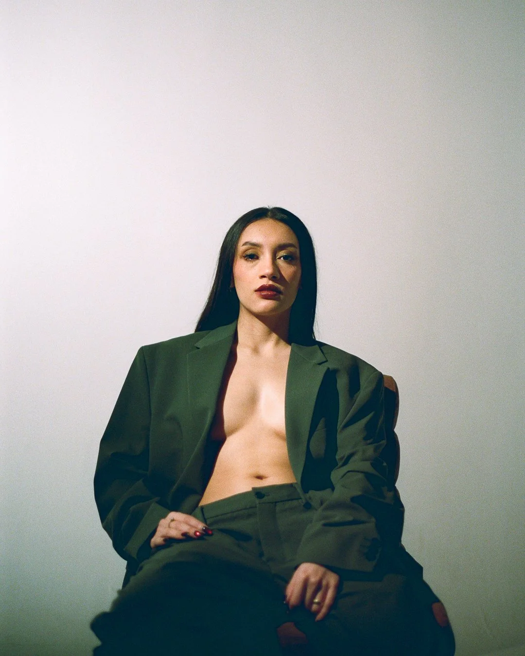 A woman with long dark hair wearing an oversized green blazer and green pants sits against a plain light-colored wall, looking directly at the camera.