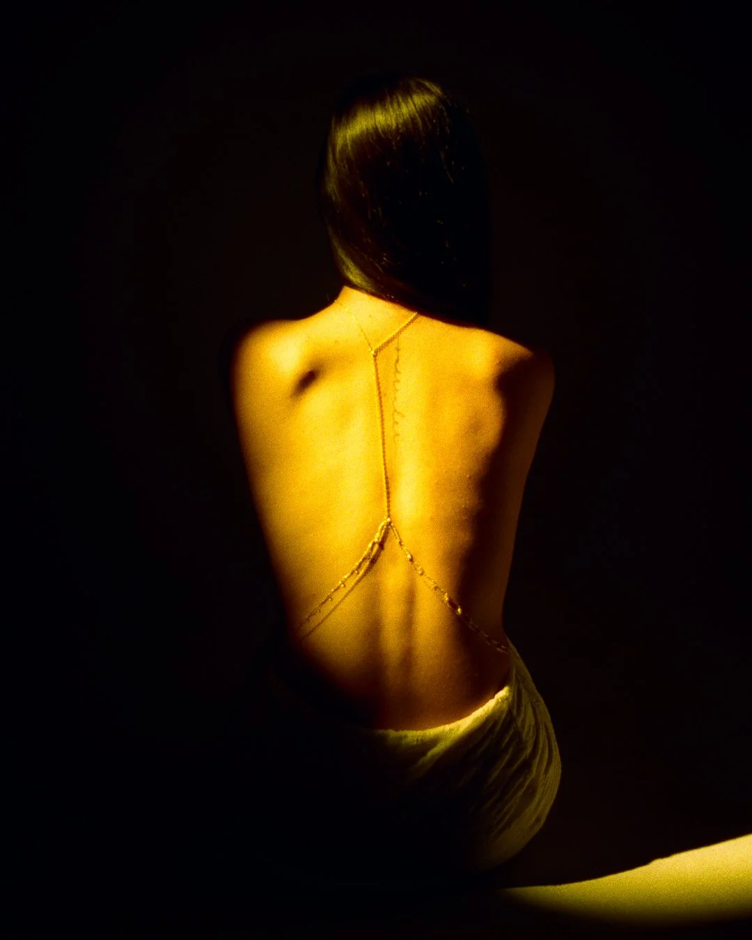 Back of a woman with long dark hair, wearing a gold body chain, in a dimly lit setting with a dark background.