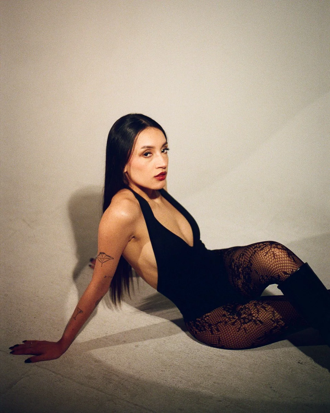 A woman with long dark hair, wearing black fishnet stockings and a revealing black outfit, sitting against a plain light-colored wall.