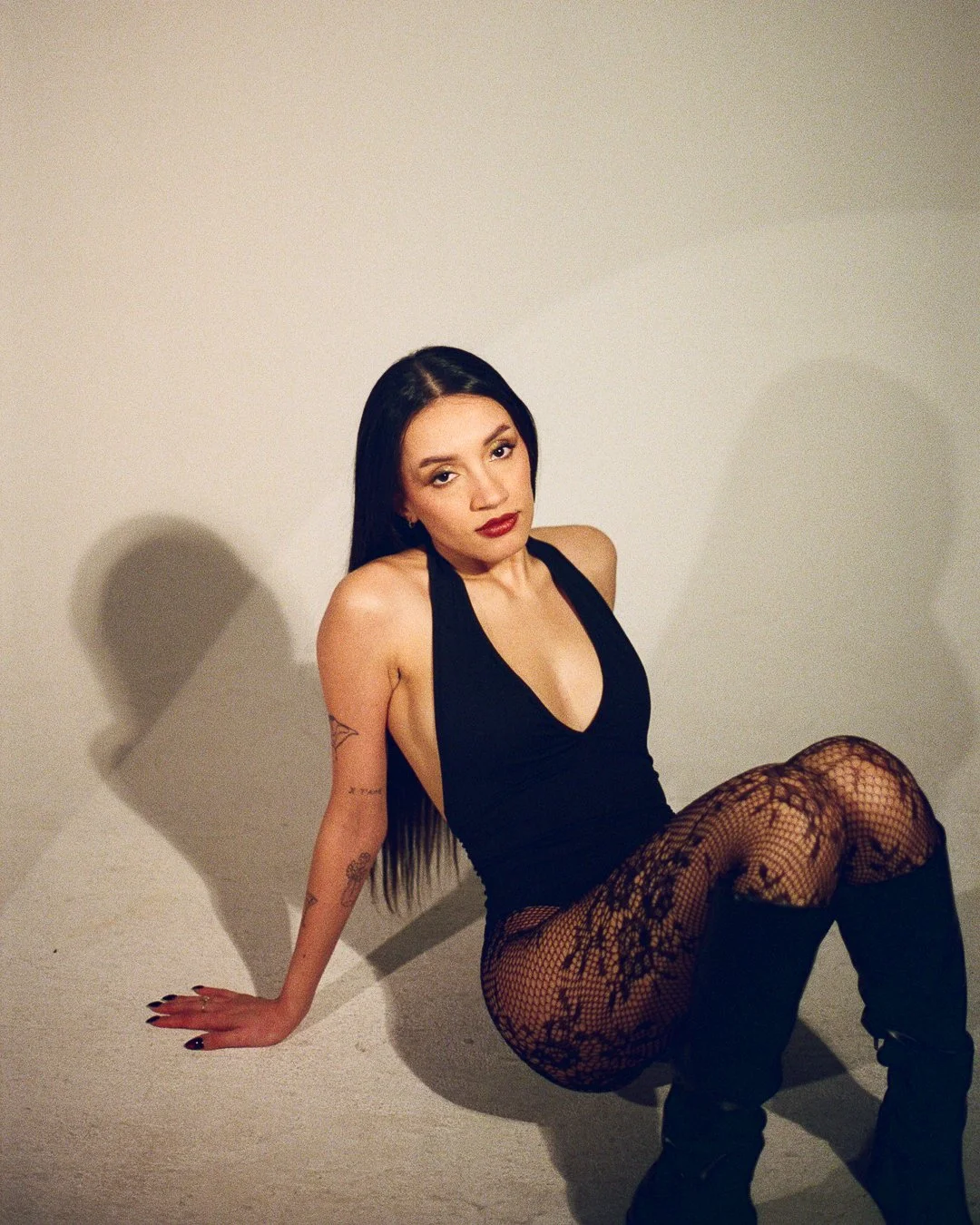A woman with long black hair, wearing a black sleeveless top, fishnet stockings, and black boots, sitting on the floor against a plain white wall, looking at the camera with a neutral expression.