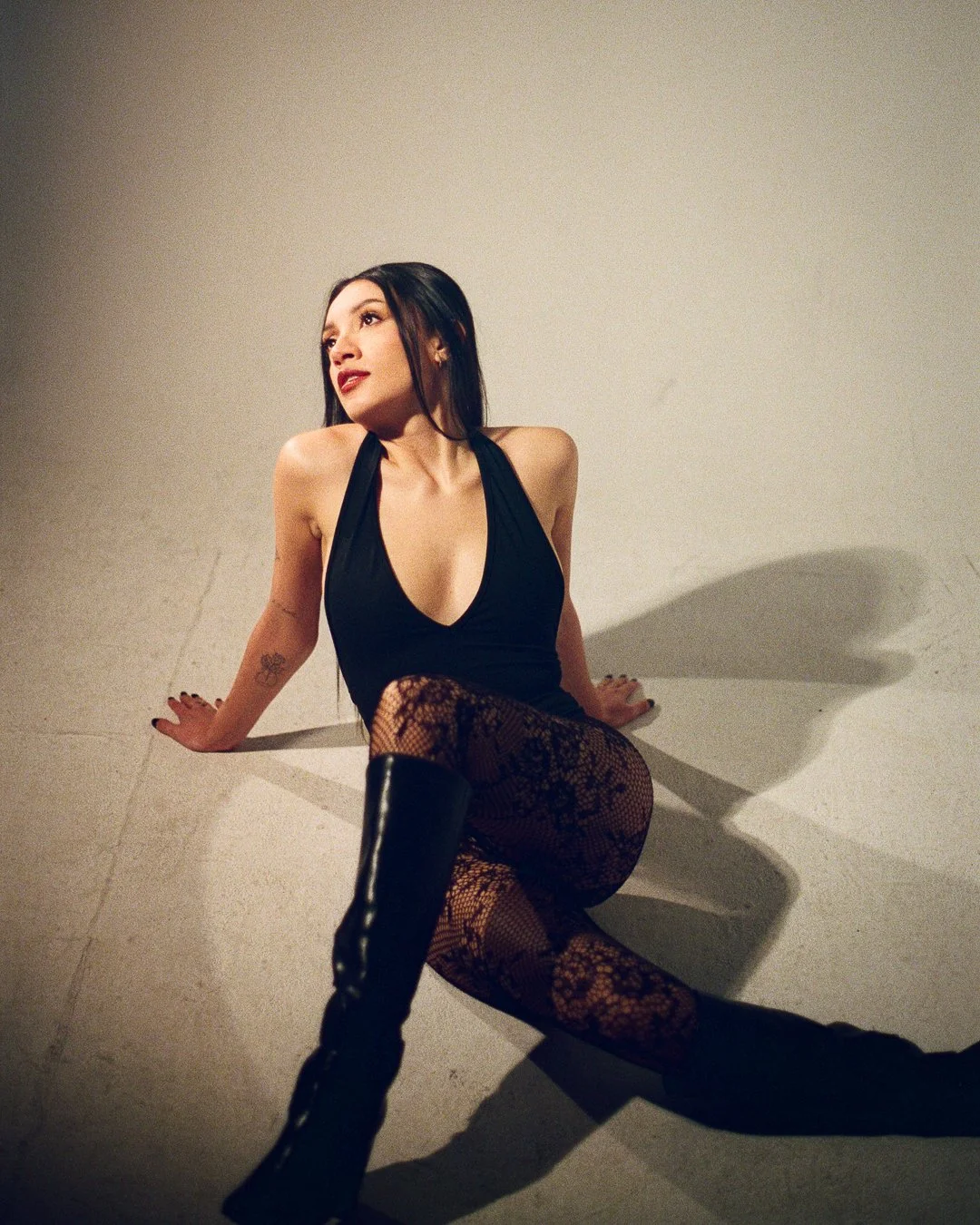 A woman in a black sleeveless top, fishnet stockings, and knee-high boots sitting on the floor against a plain wall, looking off to the side.