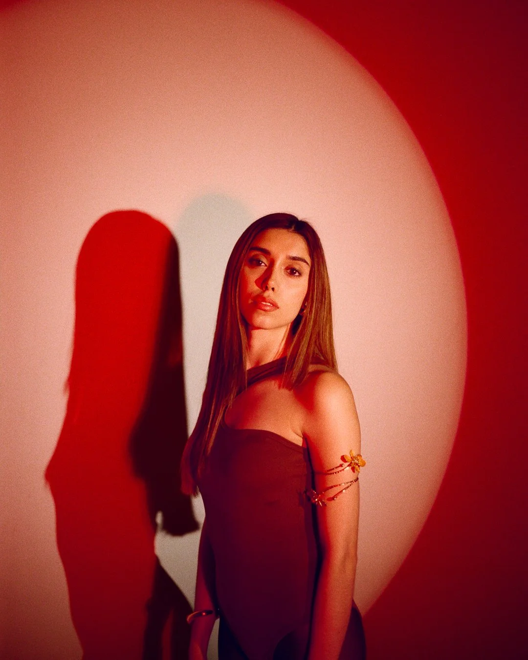 A young woman with long brown hair stands against a white wall, illuminated by red lighting, casting a shadow behind her.