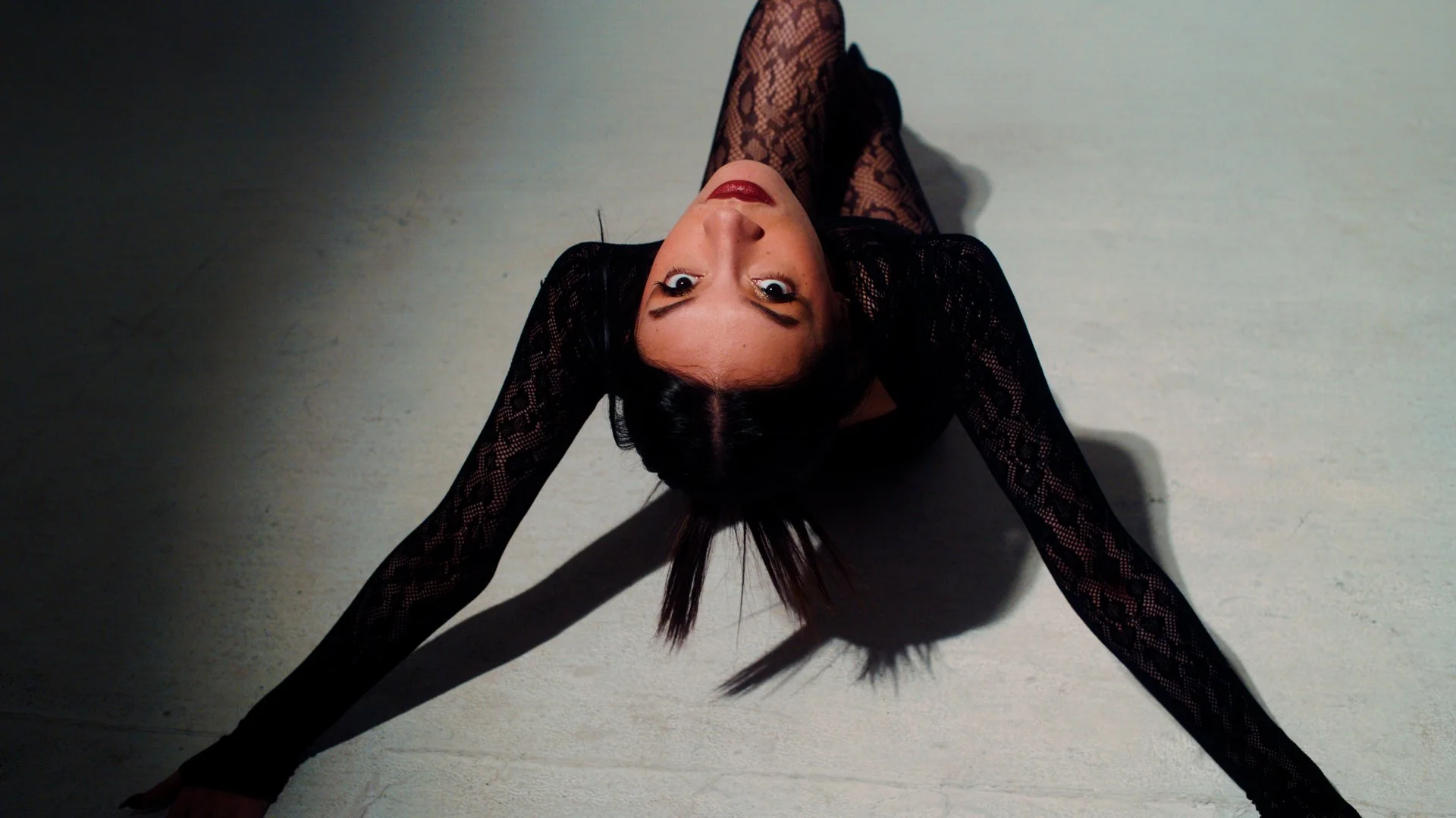 A woman with dark hair and red lipstick lies on her back on a light-colored floor, wearing a black lace dress with her arms stretched out and looking directly at the camera.