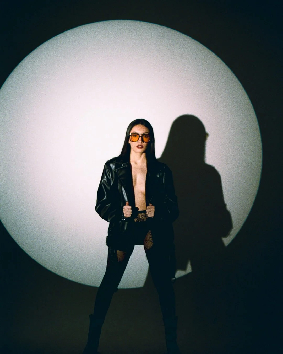 A woman with long dark hair and sunglasses standing in front of a spotlight creating a shadow on the wall behind her.