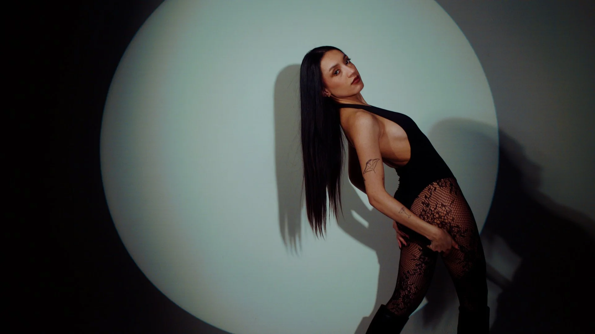 A woman with long dark hair, wearing a black bodysuit and patterned fishnet tights, stands against a plain wall with a circle of light behind her, striking a pose with her head tilted back and one hand on her thigh.