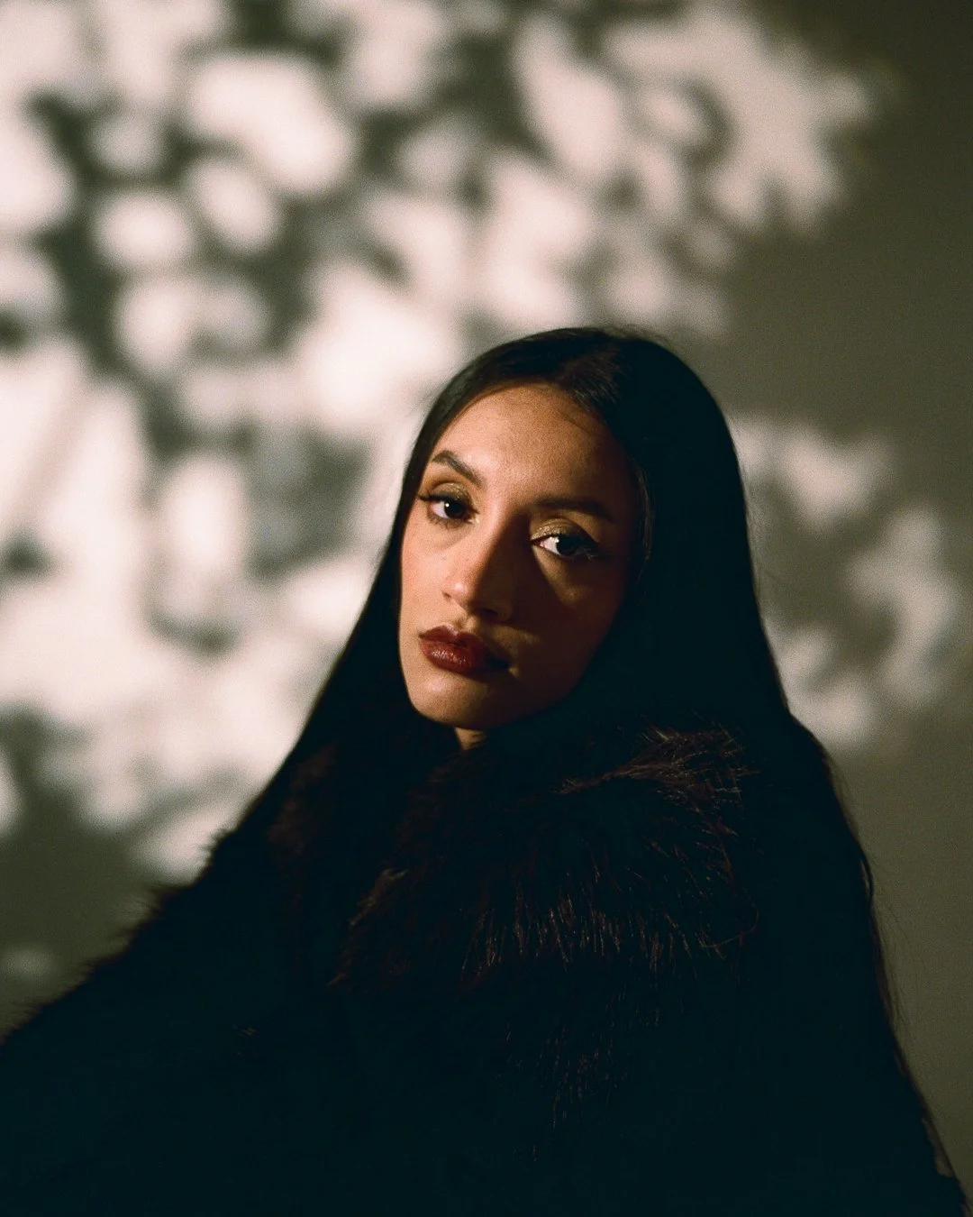 A woman with long dark hair and makeup, wearing a dark fur coat, poses indoors with soft lighting and a patterned background.