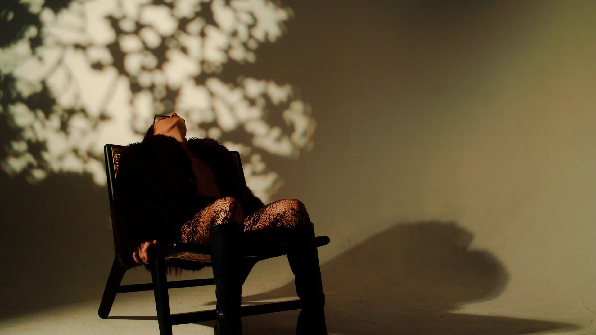 A woman sitting on a chair with her head tilted back, wearing black lace stockings and a dark fur coat, in front of a plain wall with shadows and lace-like patterns cast by a light source.