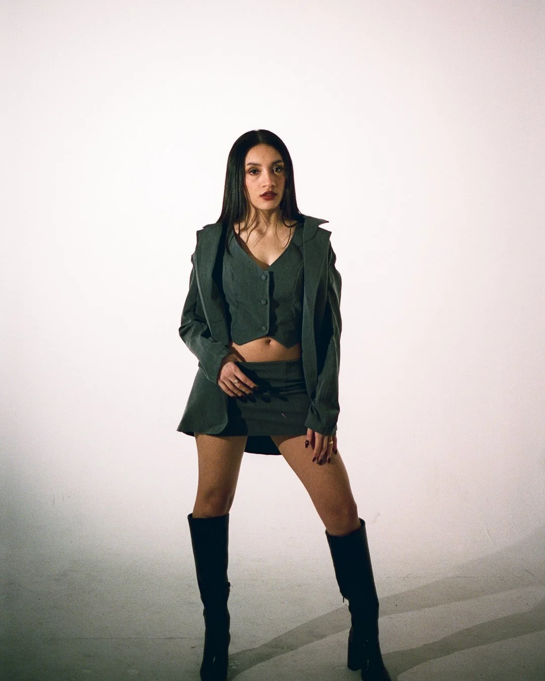 A woman standing confidently with legs apart, wearing a dark green crop top, matching skirt, oversized blazer, and tall black boots, against a plain white background.