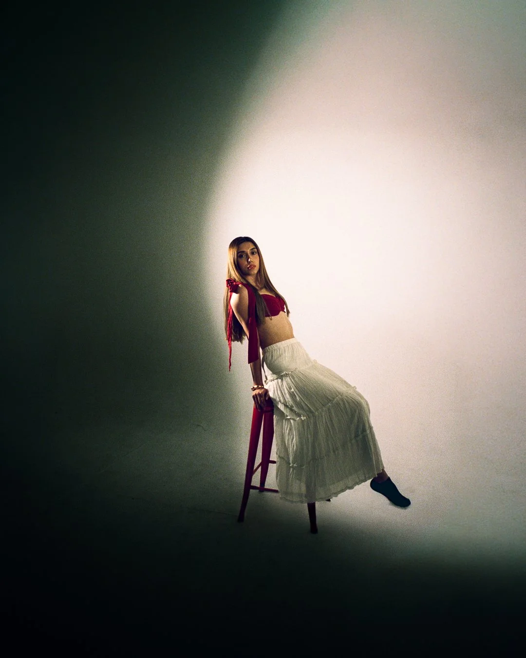 A young woman with long hair, dressed in a red crop top with tied shoulder straps and a beige pleated skirt, sitting on a red stool against a plain background with dramatic lighting.