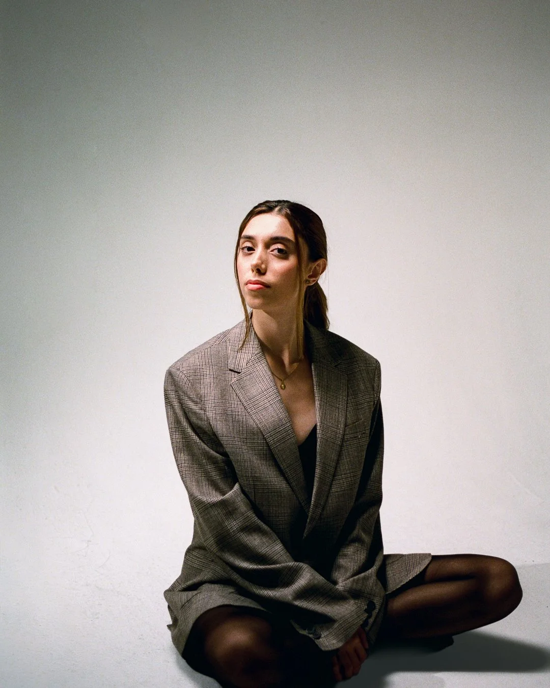 A young woman with brown hair, wearing a plaid blazer and black tights, sitting cross-legged against a plain white background, looking at the camera with a neutral expression.