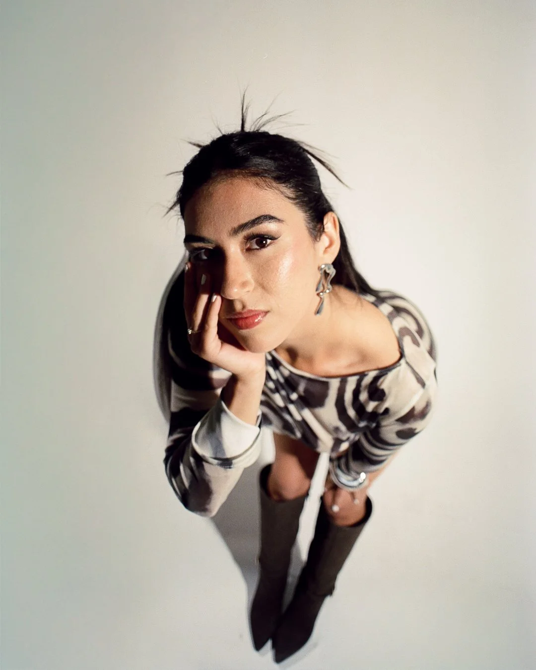 A woman with dark hair and earrings leaning forward and resting her face on her hand against a plain wall background, wearing a striped shirt.
