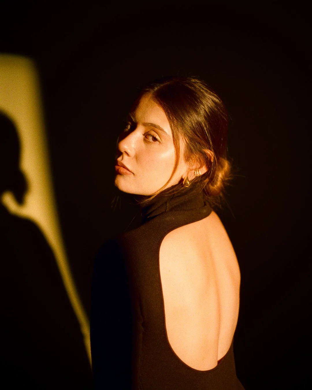 A woman with reddish-brown hair styled in a low bun, wearing a black dress with an open back, looking over her shoulder against a dark background with warm lighting.