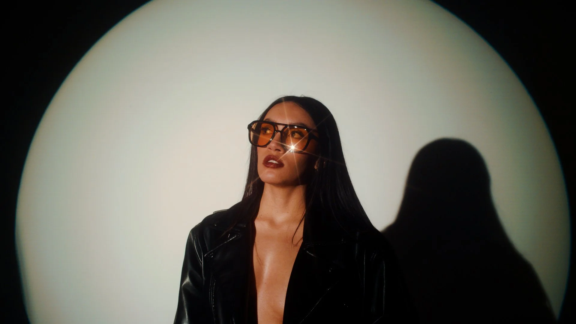 A woman with long black hair, wearing large sunglasses with orange tint, a black leather jacket, and dark lipstick, standing against a plain white background with a shadow cast behind her.