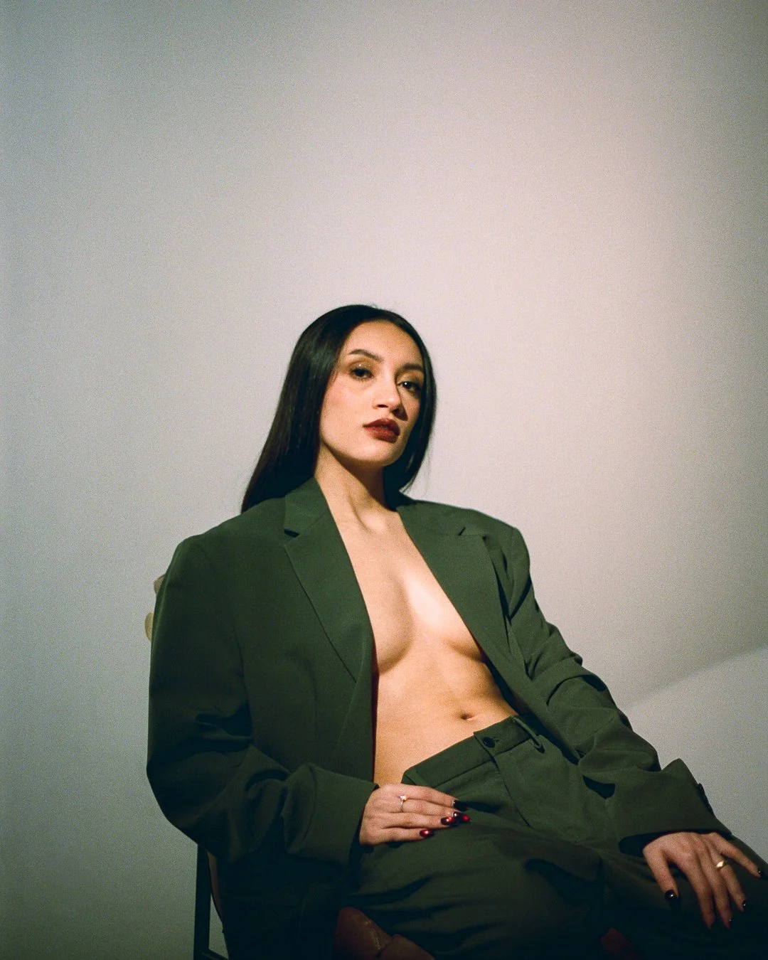 A woman with long dark hair, wearing dark lipstick and eye makeup, dressed in an oversized green blazer and matching pants, seated in front of a plain light-colored wall.