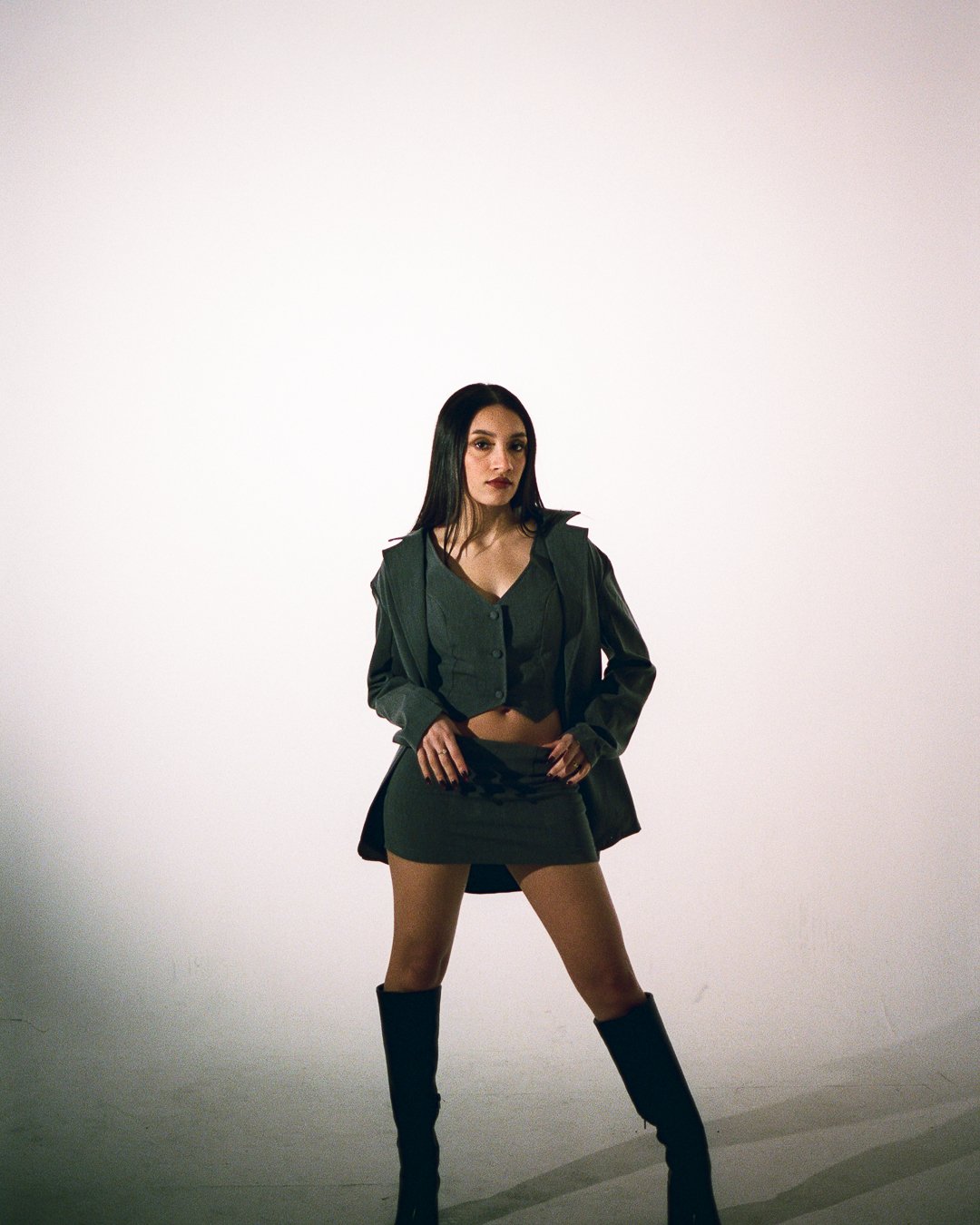 A woman with long dark hair wearing a black jacket, cropped top, mini skirt, and tall black boots standing with hands on hips against a plain white background.