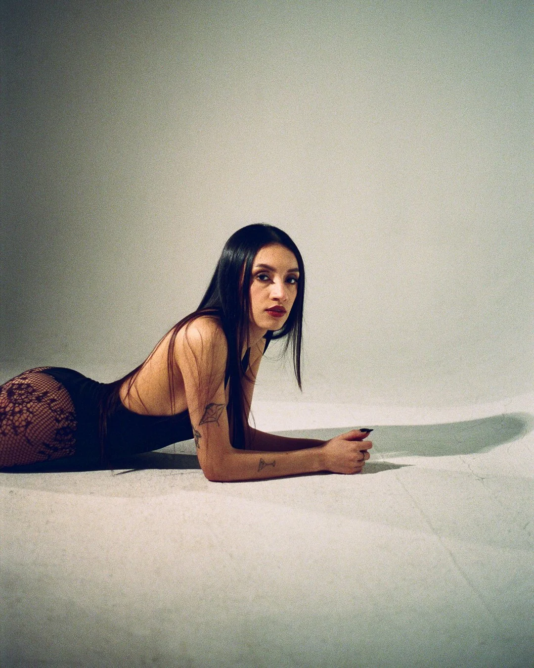 A woman with long black hair and tattoos lying on the floor, wearing a black top with lace stockings, against a plain background.