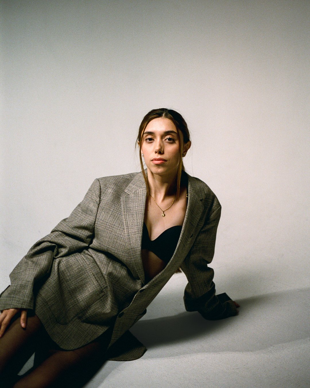 A woman wearing a grey plaid blazer and black lingerie, sitting on the floor against a plain white background.
