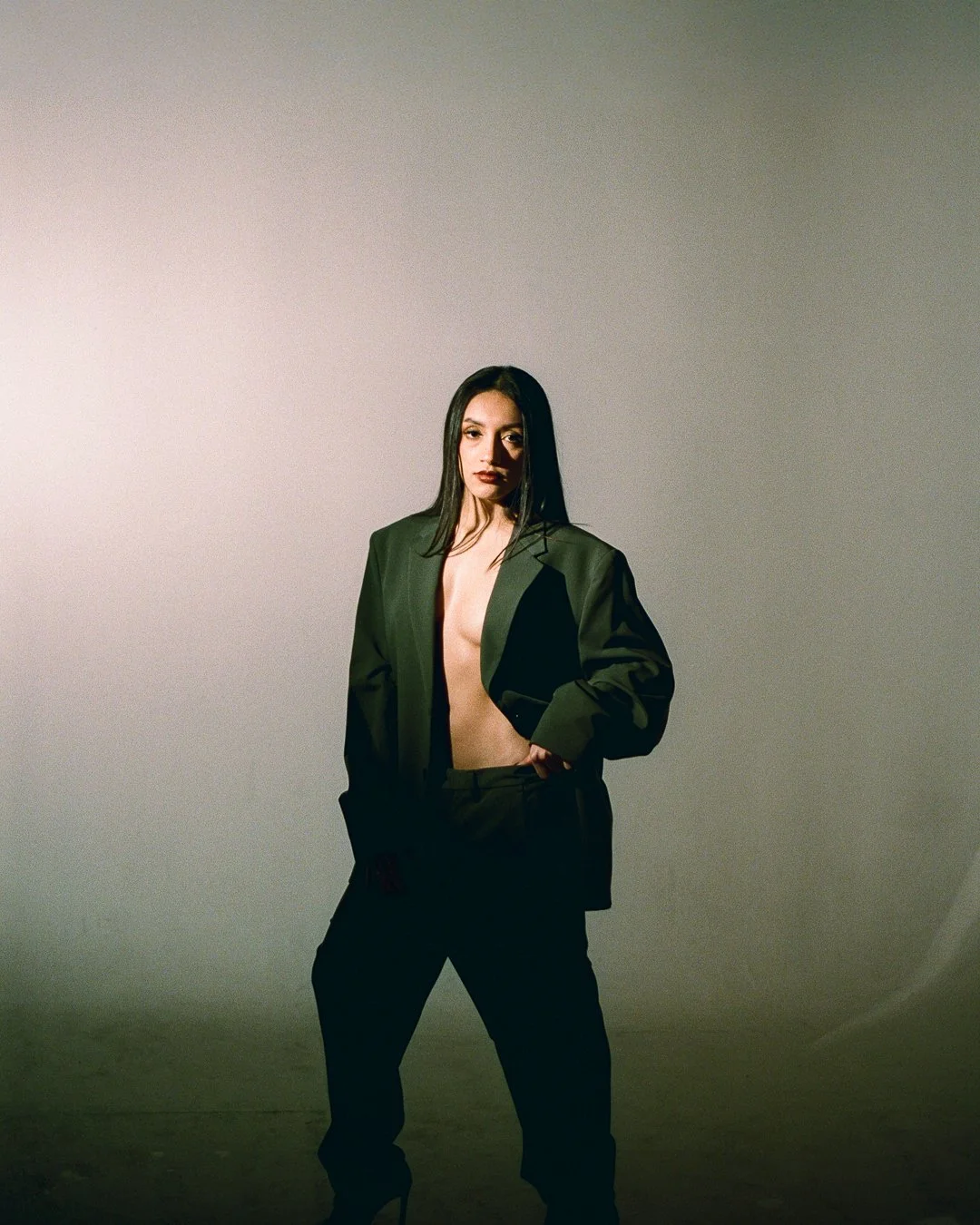 A woman with long dark hair wearing a black blazer and black pants, standing against a gradient background, with her blazer open revealing her chest.