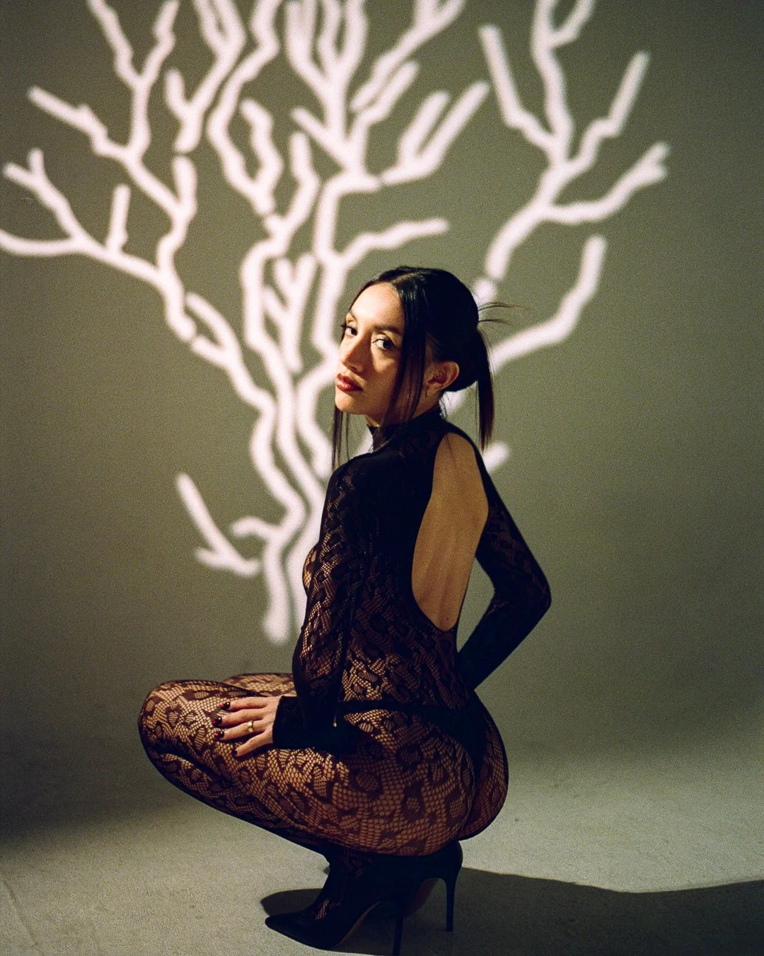 A woman dressed in black lace bodysuit with a keyhole back, wearing high heels, poses in front of a wall with a white tree branch shadow projection.