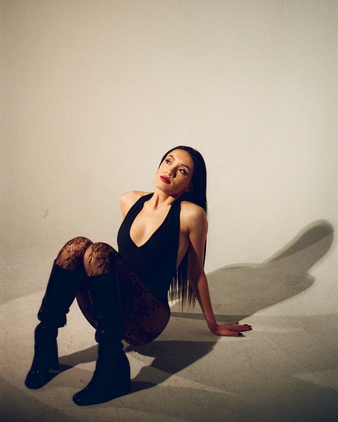 A woman with long dark hair and red lipstick, wearing a black deep V-neck sleeveless top, patterned tights, and black knee-high boots, sitting on the ground with one hand supporting her and the other arm resting on her knee, against a plain white bac