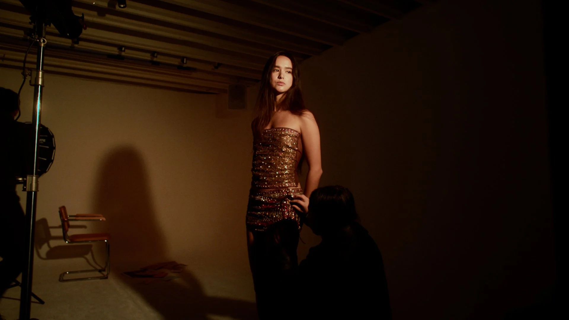 A woman in a shiny, sequin dress stands in front of a plain background during a photo shoot, with a resting chair and a person crouching in front of her.