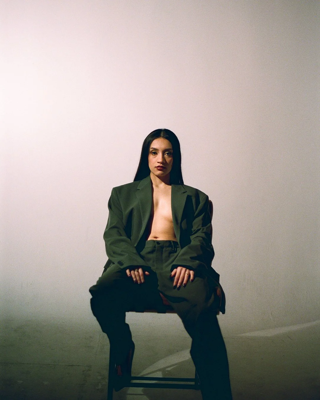 A woman with long dark hair, wearing a green blazer and pants, sits on a chair against a plain white wall, looking directly at the camera.